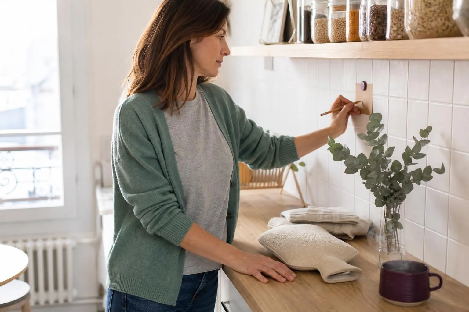 Femme d’environ quarante ans debout dans une cuisine claire, notant quelques mots au crayon sur une fiche aimantée posée près d’une bouilloire